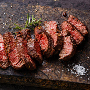 Slices of medium-rare steak arranged on a dark wooden board with a rosemary sprig and flaky salt.